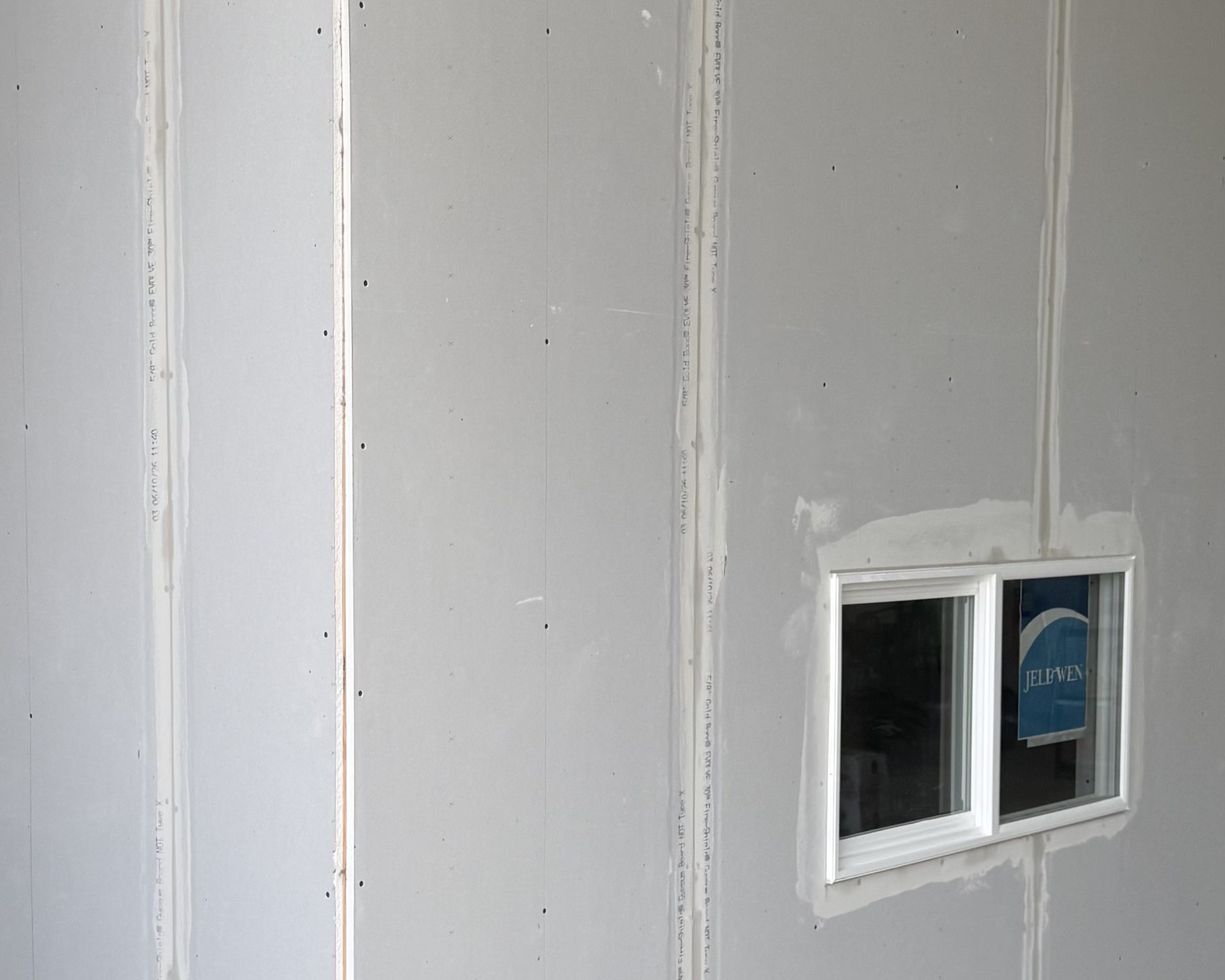 Drywall hung on a bedroom wall, seams set with tape and the screw heads still showing, around a small white sliding window.